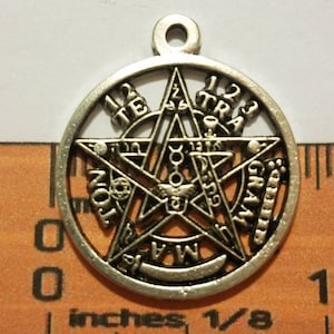 May include: A silver-toned metal circular pendant featuring a central pentagram design with the text TETRAGRAMMATON embossed around the perimeter. The pendant is positioned against a ruler showing a diameter of approximately 1.25 inches.