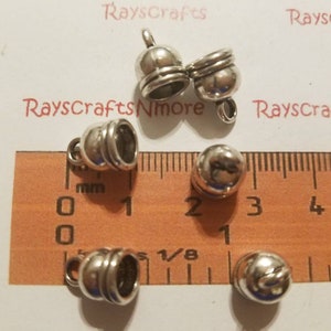 May include: Several small, silver-colored metal bell-shaped beads with a loop at the top for stringing. A ruler with millimeter markings is next to the beads. The beads are approximately 0.5 inches in diameter.