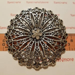 Puede incluir: Un broche plateado de estilo filigrana con un grupo central de pedrería transparente. El broche tiene forma circular con intrincados patrones calados que irradian hacia afuera. Una regla de madera se coloca junto al broche, mostrando medidas en centímetros.