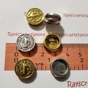 May include: A collection of metallic Saint Benedict round charms in gold and silver tones. The charms feature detailed religious imagery and are displayed with a ruler for size reference. The text "Rayscrafts Nmore" is visible.