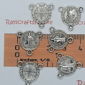 May include: Five silver-colored Saint Benedict medal connectors. Each connector features a round design with a saint or cross image, and three small loops for attaching to other components. The connectors are displayed on a wooden ruler.