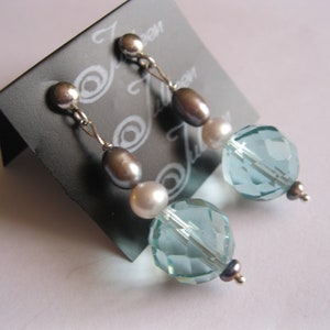 May include: Dangle earrings featuring a silver post, a gray oval bead, a white round bead, and a faceted light blue crystal bead. The earrings are displayed on a black card with the text "Jeen".