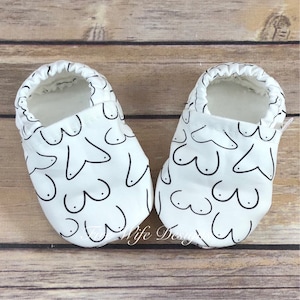 May include: A pair of white baby booties with a black outline of a breast pattern.