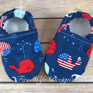 May include: A pair of blue baby booties with a whale print. The whales are red, white, and blue and have an American flag design. The booties are lined with white fleece.