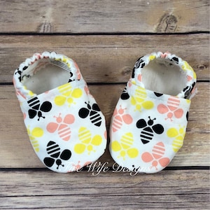 May include: A pair of white baby booties with a bee pattern. The bees are black, yellow, and pink. The booties are soft and comfortable.