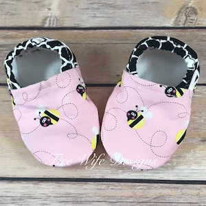 May include: A pair of pink baby booties with a bee pattern. The booties have a black and white trim around the top.
