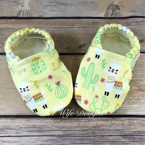 May include: A pair of yellow baby booties with a llama and cactus print. The booties are soft and comfortable, perfect for keeping little feet warm.