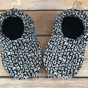 May include: A pair of black baby booties with a white alphabet print. The booties are made of soft fabric and have a comfortable fit.