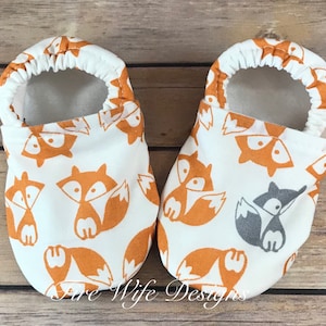 May include: A pair of white baby booties with a fox print. The booties have a soft, white lining and a brown elastic band around the top.