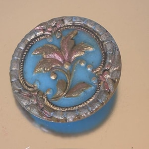 May include: A round, antique button with a light blue center and a detailed floral design. The floral design features gold and pink accents. The button is framed by a silver-colored, textured border.