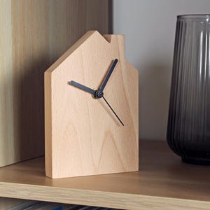May include: A wooden house-shaped clock with black clock hands. The clock is made of light-colored wood and sits on a wooden shelf. A dark gray glass vase is to the right of the clock. The clock is a decorative home decor item.