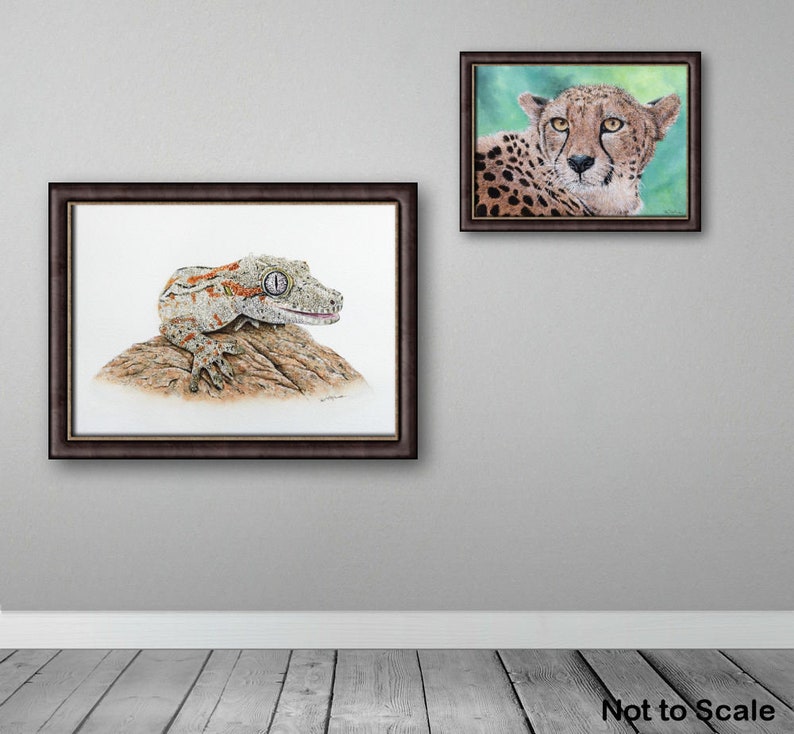 In this photo the gecko is shown in a brown frame and is hung on a wall alongside a painting of a cheetah in the same type of frame.