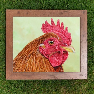 The rooster painting in a wooden frame laid on a closely cut lawn.