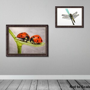 In this photo the ladybird painting is shown in a dark frame, hung on a grey wall.  There is also a painting of a dragonfly, slightly smaller and to the right.