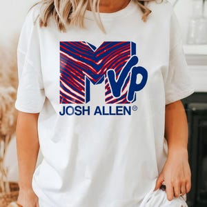 May include: White t-shirt featuring a blue and red graphic design with the letters "MVP" and the name "JOSH ALLEN" in blue. The "M" and "V" are in a red and blue pattern. The shirt is a casual style.