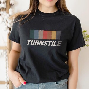 May include: Black t-shirt featuring the word "TURNSTILE" in white, with a rectangular color block design above the text. The color block includes shades of green, blue, gray, red, yellow, and brown.