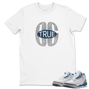 Op de afbeelding: Een wit T-shirt met korte mouwen en een grote afbeelding op de borst met de letters GG in een zwart-grijs olifantenpatroon, met een blauwe ovaal met het woord TRUE in witte letters, gecombineerd met wit-blauwe sneakers.