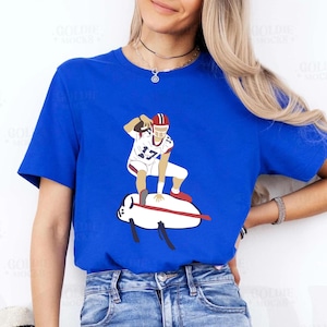 May include: Royal blue t-shirt featuring a graphic of a football player in a white uniform with red accents, holding a football. The player is standing on a white and red object. The shirt is a solid color.