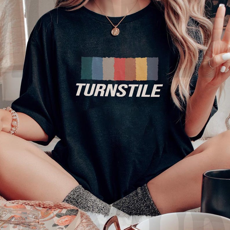 May include: Black t-shirt with the word "TURNSTILE" in white block letters. Above the word is a horizontal rectangle divided into six colored sections: green, blue, gray, red, orange, and yellow. The shirt is a casual style.