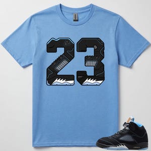 Op de afbeelding: Een lichtblauw t-shirt met korte mouwen en een grote afbeelding van het getal 23 in het zwart, ontworpen met texturen geïnspireerd op sneakers en lichtblauwe accenten. Een zwarte en lichtblauwe hoge sneaker staat in de rechterbenedenhoek.