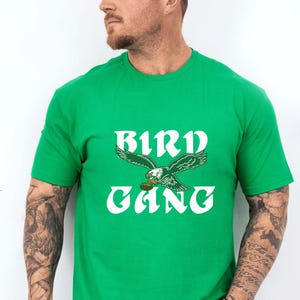 Può includere: Una maglietta verde con la scritta bianca "BIRD GANG" a forma di arco. Un'aquila verde e bianca è raffigurata sopra la parola "GANG". La maglietta è a maniche corte.
