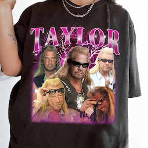 May include: Dark gray t-shirt featuring a collage of images with a man wearing sunglasses. The word "TAYLOR" is printed in large, pink, retro-style lettering above the images. The background has a purple and white lightning bolt design.