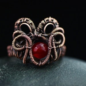 May include: A handcrafted copper wire ring with a central red gemstone. The ring features intricate wire wrapping in a floral design. The gemstone is a round, faceted, and vibrant red colour. The ring is set against a dark background.