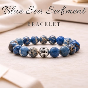 May include: A beaded bracelet featuring round blue sea sediment jasper stones with varied marbling and a single central silver-toned metal bead with a textured, ribbed design. The text Blue Sea Sediment BRACELET is displayed above the jewelry.