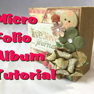 Op de afbeelding: Een klein, bruin, met stof bekleed album met een bloemenmotief. Het album heeft de woorden "live.love.journal" op de voorkant gedrukt. Het album is versierd met bloemen, knopen en glitter.