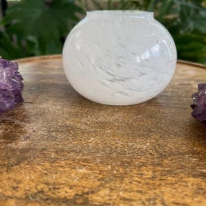 May include: A round, white glass vase with a speckled design. The vase sits on a wooden surface, with purple amethyst crystals on either side. The vase has a wide opening and a spherical shape.