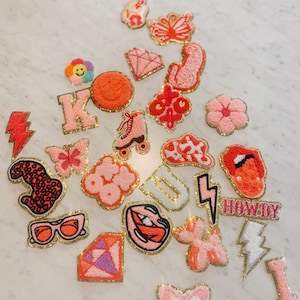 May include: A collection of colorful embroidered iron-on patches featuring various designs, including a roller skate, a flower, a lightning bolt, a butterfly, a diamond, a smiley face, a letter K, a letter U, a mouth, and the word "HOWDY".