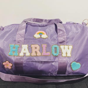 May include: Purple duffel bag with a rainbow, flower, and heart applique. The bag has the name "HARLOW" spelled out in colourful, glittery letters.