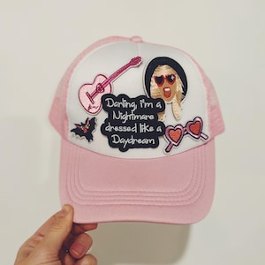 Può includere: Cappellino da camionista rosa e bianco con varie toppe ricamate, tra cui una chitarra, una donna con occhiali da sole, un pipistrello e cuori. Il testo sul cappello recita "Darling, I'm a Nightmare dressed like a Daydream."