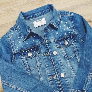 May include: A blue denim jacket with pearl embellishments on the shoulders and pockets. The jacket has a button closure and two front pockets. The label on the jacket reads "Old Navy".