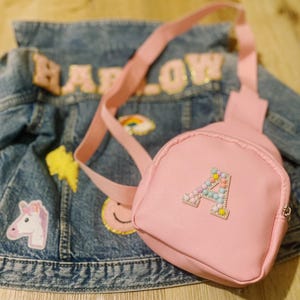 Puede incluir: Una mochila rosa de imitaci&oacute;n de cuero con una letra A de cuentas en la parte delantera. La mochila est&aacute; sobre una chaqueta vaquera con parches.