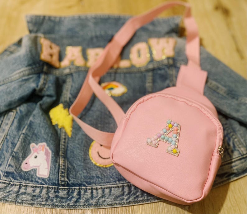 Puede incluir: Una mochila rosa de imitaci&oacute;n de cuero con una letra A de cuentas en la parte delantera. La mochila est&aacute; sobre una chaqueta vaquera con parches.