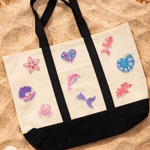 May include: Cream tote bag with black handles and base, decorated with embroidered patches of sea creatures. Patches include a starfish, heart, seashell, dolphin, mermaid tail, crab, and coral. The bag is on sand.