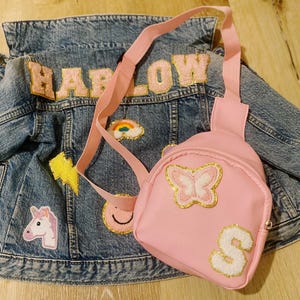 Puede incluir: Una chaqueta vaquera rosa con el nombre "HARLOW" en letras de purpurina rosa. La chaqueta tiene varios parches, incluyendo un arco&iacute;ris, un rayo, una carita sonriente y un unicornio. Una peque&ntilde;a mochila rosa con una mariposa y la letra "S" en purpurina est&aacute; unida a la chaqueta.