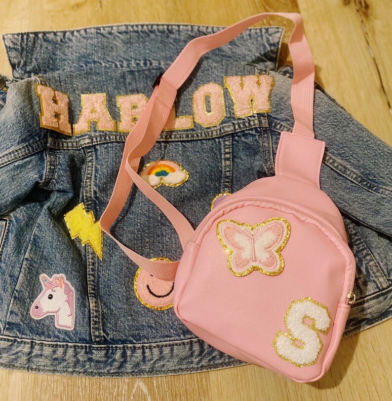 Puede incluir: Una chaqueta vaquera rosa con el nombre "HARLOW" en letras de purpurina rosa. La chaqueta tiene varios parches, incluyendo un arco&iacute;ris, un rayo, una carita sonriente y un unicornio. Una peque&ntilde;a mochila rosa con una mariposa y la letra "S" en purpurina est&aacute; unida a la chaqueta.