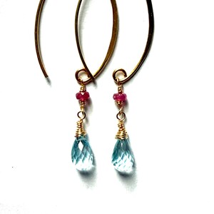Peut inclure: Une paire de boucles d'oreilles dorées en forme de goutte d'eau. Chaque boucle d'oreille comporte un petit groupe de perles rouges et une pierre précieuse en forme de goutte d'eau, bleu clair et facettée. Les boucles d'oreilles sont présentées sur fond blanc.