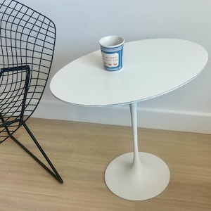 May include: A white oval-shaped side table with a single stem and circular base. A small blue and white cup with text sits on the table. A black wire chair is in the background.