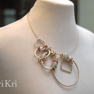 May include: Gold necklace with a geometric design featuring multiple interconnected circles and a square. The circles are decorated with pink, green, and gold accents.