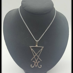 Könnte beinhalten: Silberne Kettenkette mit einem silbernen Anhänger mit dem Siegel des Dämons Baphomet. Der Anhänger ist ein Pentagramm mit einem Kreuz im Inneren und zwei Hörnern unten. Die Kette liegt auf einem schwarzen Präsentationsständer mit dem Schriftzug "Poison Kiss" auf der Basis.