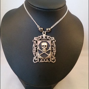 Puede incluir: Un collar de plata con un colgante de calavera y huesos cruzados en negro y plata. El colgante está enmarcado en un diseño de filigrana de plata. El collar tiene tres cuentas negras.