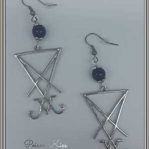 Op de afbeelding: Een paar zilveren oorbellen met een zwarte kraal en een zilveren hanger in de vorm van een pentagram met een kruis erin. De oorbellen zijn gelabeld met "Poison Kiss".