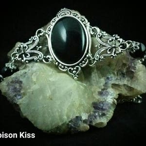 Könnte beinhalten: Ein silbernes Armband mit einem großen, schwarzen, ovalen Edelstein, der in einem filigranen Rahmen gefasst ist. Das Armband ist mit kleineren schwarzen Edelsteinen und filigranen Blumenmustern verziert.
