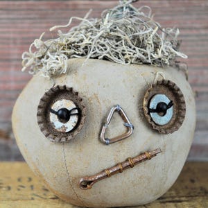 May include: A handcrafted, whimsical face made from a light tan fabric. Bottle caps form the eyes, a metal triangle is the nose, and a copper rod with wire wraps the mouth. The head is topped with dried, light-colored plant material.