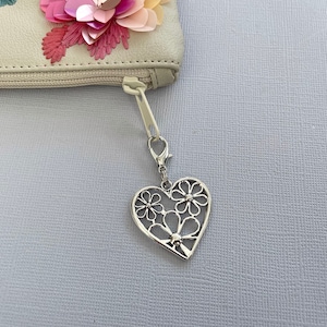 May include: A silver heart-shaped zipper pull with a delicate floral design. The pull is attached to a white zipper.