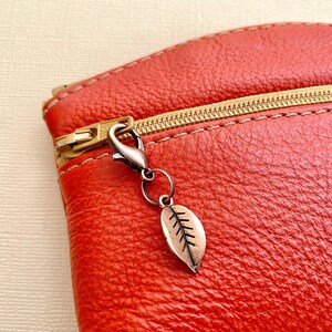 May include: A close-up of a rust-coloured leather pouch with a gold zip and a leaf charm. The charm is a small, detailed leaf pendant with a bronze finish, attached to a silver-coloured clasp. The leather has a textured appearance.