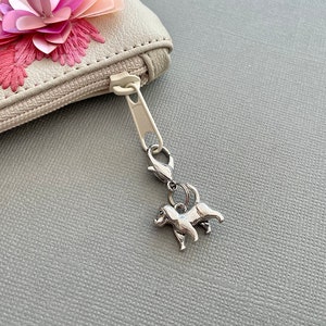 May include: A silver dog charm hanging from a zip. The charm is a small, detailed figurine of a dog. The zip is attached to a light-coloured pouch with a floral decoration.